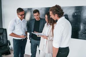 com documentos em mãos. grupo de jovens inteligentes que estão perto do quadro-negro no escritório foto