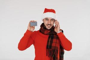 com telefone e cartão de crédito. jovem bonito em roupas de ano novo em pé dentro de casa contra um fundo branco foto