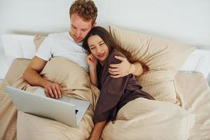 casal deitado na cama com o laptop. interior e design do lindo quarto moderno durante o dia foto