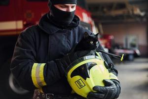 retrato de bombeiro em uniforme de proteção que segura um lindo gatinho preto foto