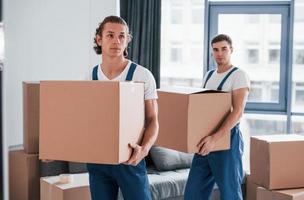 com caixas nas mãos. dois jovens em uniforme azul trabalhando dentro de casa na sala foto