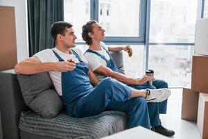 sentado no sofá. cansado e descansando. dois jovens em uniforme azul trabalhando dentro de casa na sala foto