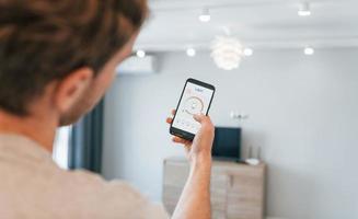 segura o telefone. visão traseira do homem adulto que está dentro de casa controlando a tecnologia de casa inteligente foto