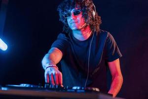 homem com cabelo encaracolado usando equipamento de dj e parado na sala escura iluminada por neon foto