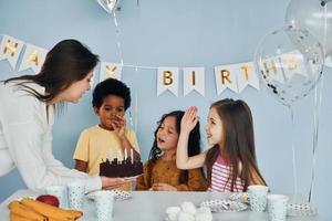 mulher segura bolo. as crianças comemorando a festa de aniversário dentro de casa se divertem juntas foto