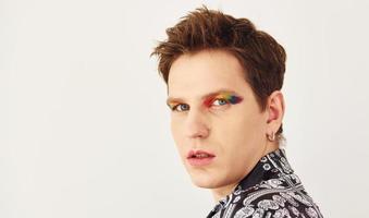 jovem gay está de pé no estúdio e posando para uma câmera. maquiagem multicolorida nos dedos do rosto foto