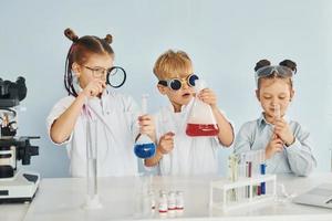 de pé ao lado da mesa com tubos de ensaio. crianças em jalecos brancos interpretam cientistas em laboratório usando equipamentos foto