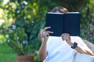 mulher asiática segurando um livro na frente do rosto enquanto está sentado e lendo o canto do banco do parque. conceito. mulher asiática fazendo atividades ao ar livre, como ler livros, trabalhar, fazer um piquenique com a família foto