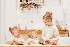 3 loiras decoram biscoitos de natal com glacê em uma mesa de madeira na cozinha. as namoradas estão cozinhando juntas na cozinha com decoração de natal. foto