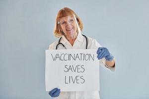 vacinação salva bandeira de vidas. médica sênior de jaleco branco está de pé dentro de casa foto