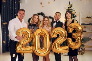 segurando balões com o número 2023. grupo de pessoas tem uma festa de ano novo dentro de casa juntos