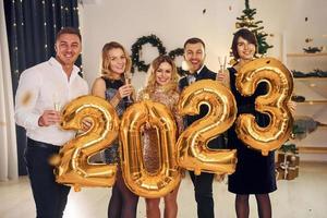 segurando balões com o número 2023. grupo de pessoas tem uma festa de ano novo dentro de casa juntos