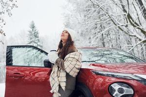 cena não urbana. bela jovem está ao ar livre perto de seu automóvel vermelho no inverno foto