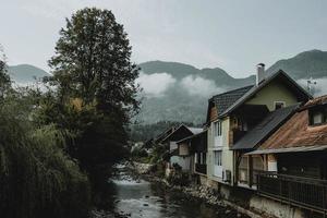 rio de montanha e fileira de casas perto dele na cidade eslovena bohinjska bistrica sob os Alpes Julianos escondido nas nuvens ao fundo durante o nascer do sol durante a manhã de verão foto