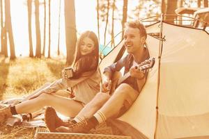 cara tocando violão. jovem casal está viajando na floresta durante o dia juntos foto