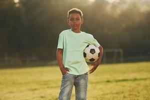 senta-se com bola de futebol. garoto afro-americano divirta-se no campo durante o dia de verão foto