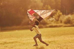 correndo com pipa vermelha. garoto afro-americano divirta-se no campo durante o dia de verão foto