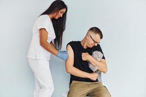 homem segurando ursinho de pelúcia durante o procedimento. médico de uniforme fazendo vacinação para o paciente foto