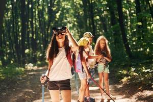garota de pé na frente de seus amigos. crianças passeando na floresta com equipamentos de viagem foto