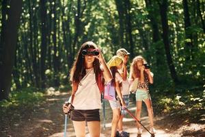 garota de pé na frente de seus amigos. crianças passeando na floresta com equipamentos de viagem foto