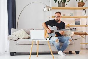 usando laptop. homem em roupas casuais e com violão está dentro de casa foto