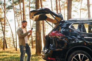 perto do carro. homem de jeans está ao ar livre na floresta com seu automóvel preto foto