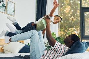 deitada na cama e se divertindo. pai afro-americano com seu filho em casa foto