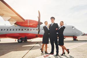 piloto e duas aeromoças. tripulação de trabalhadores do aeroporto e avião em roupas formais juntos ao ar livre foto