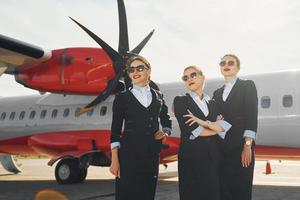 três aeromoças. tripulação de trabalhadores do aeroporto e avião em roupas formais juntos ao ar livre foto