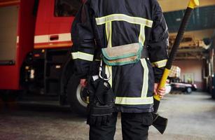 detém o machado nas mãos. bombeiro masculino em uniforme de proteção em pé perto do caminhão foto