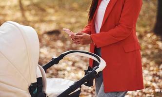 com telefone na mão. mãe de casaco vermelho dá um passeio com seu filho no carrinho no parque no outono foto