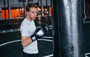 jovem de camisa branca e luvas de proteção de boxe fazendo exercícios no ginásio com saco de empurrar foto