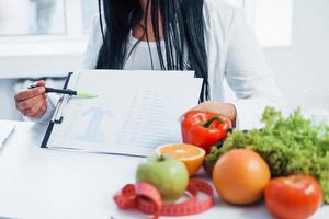 nutricionista feminina em jaleco branco sentado dentro de casa no escritório no local de trabalho e mostrando informações no documento foto