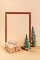composição mínima festiva com moldura de madeira vazia e árvores de natal de madeira e caixa de presente de papelão em bege. foto