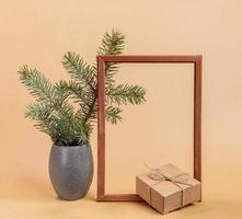composição mínima da moda festiva com galhos de árvores de abeto em vaso cinza, moldura de madeira vazia, caixa de presente artesanal em bege. foto