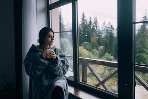 mulher segurando caneca, sentada no parapeito da janela e olhando para as montanhas foto