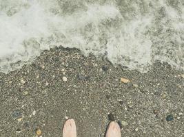 praia de seixos com pedras. um cara de sapatos está parado na praia. resto dos turistas no mar. os pés são lavados pelas ondas do mar, espuma branca foto