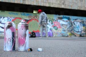 várias latas de spray usadas com tinta rosa e branca estão no asfalto contra o cara de pé em frente a uma parede pintada em desenhos de graffiti coloridos foto