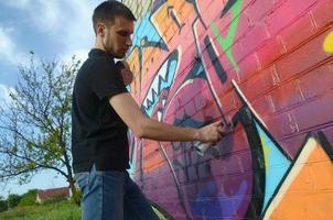 jovem grafiteiro com mochila e máscara de gás no pescoço pinta graffiti colorido em tons de rosa na parede de tijolos. arte de rua e processo de pintura contemporânea foto