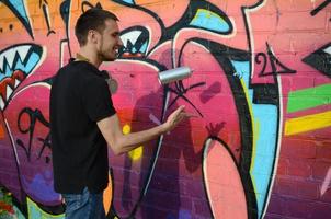 jovem grafiteiro com máscara de gás no pescoço joga sua lata de spray contra graffiti rosa colorido na parede de tijolos. arte de rua e processo de pintura contemporânea foto