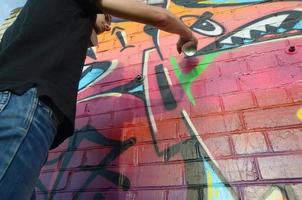 jovem grafiteiro com mochila e máscara de gás no pescoço pinta graffiti colorido em tons de rosa na parede de tijolos. arte de rua e processo de pintura contemporânea foto