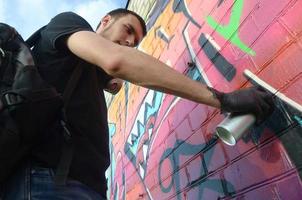 jovem grafiteiro com mochila e máscara de gás no pescoço pinta graffiti colorido em tons de rosa na parede de tijolos. arte de rua e processo de pintura contemporânea foto