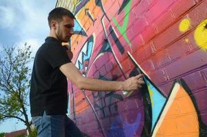 jovem grafiteiro com mochila e máscara de gás no pescoço pinta graffiti colorido em tons de rosa na parede de tijolos. arte de rua e processo de pintura contemporânea foto