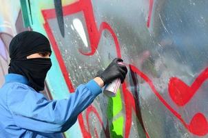 um jovem hooligan com um rosto escondido pinta graffiti em uma parede de metal. conceito de vandalismo ilegal foto