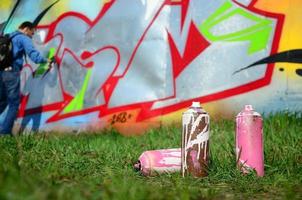 algumas latas de tinta usadas contra o fundo do espaço com a parede na qual o jovem faz um grande desenho de grafite. arte moderna de desenhar paredes em graffiti foto