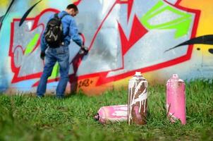 algumas latas de tinta usadas contra o fundo do espaço com a parede na qual o jovem faz um grande desenho de grafite. arte moderna de desenhar paredes em graffiti foto