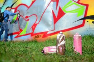 algumas latas de tinta usadas contra o fundo do espaço com a parede na qual o jovem faz um grande desenho de grafite. arte moderna de desenhar paredes em graffiti foto