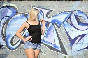 garota loira caucasiana sexy em shorts jeans e blusa preta posando contra a parede de graffiti durante o dia ao ar livre foto