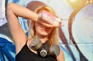 jovem e bela garota sexy grafiteiro com máscara de gás no pescoço, escondendo os olhos com uma lata de spray em pé sobre um fundo de parede com um padrão de graffiti em tons de azul e roxo foto