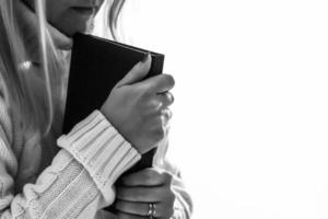 uma mulher está segurando e abraçando a bíblia no peito pela manhã. bíblia é capa preta. fundo cinza e branco. foto